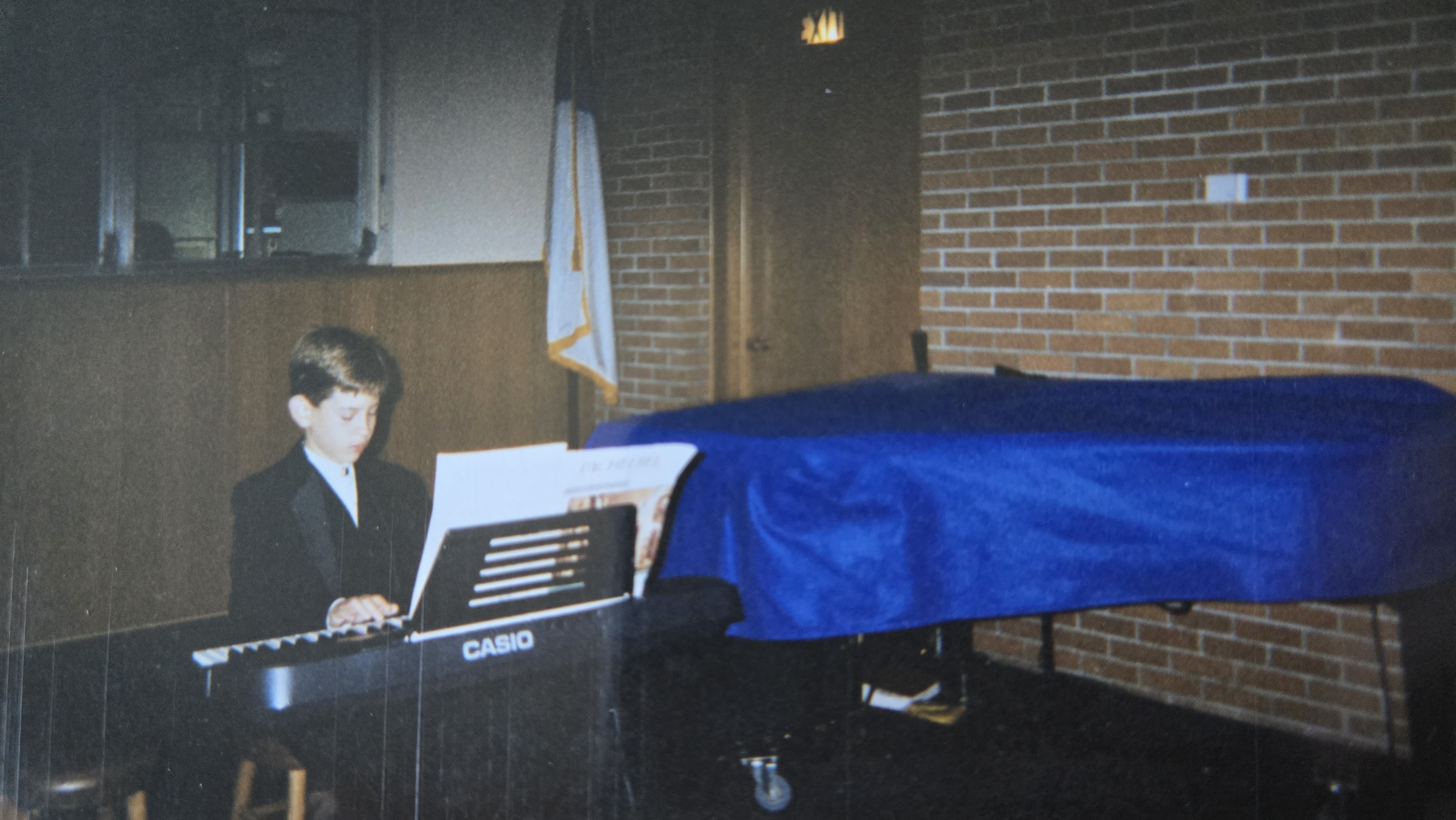 A disposable camera photograph of a young boy in a tuxedo sitting at a Casio keyboard.